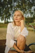 A young woman with blonde hair rests her chin on her hand while sitting on a bench in a park.