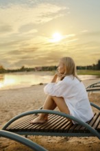 A woman sits pensively on a wooden lounge chair by the lakeside as the sun sets. She gazes at the
