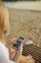 A woman sits on a wooden lounge, absorbed in her smartphone. The sun sets in the background,