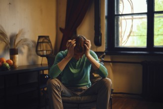 A man sits on a chair visibly overwhelmed and stressed. He put his head in his hands in frustration