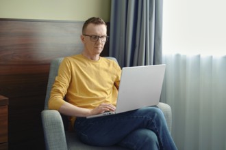 A man sits in a comfortable chair, focused on his laptop in a bright room. Soft natural light