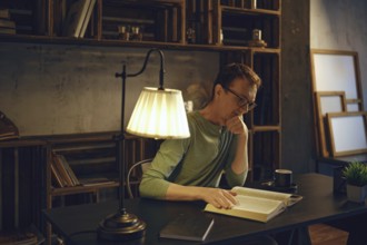 A man sits at a desk in a dimly lit room, engrossed in a book. Warm light from a lamp highlights