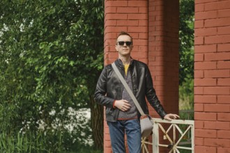 A man stands with a relaxed posture beside vibrant red brick pillars, dressed in a casual outfit