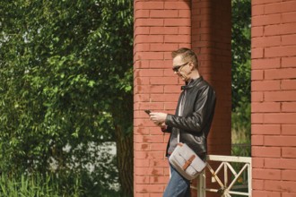 A man wearing sunglasses and a leather jacket stands by red brick columns, attentively using his