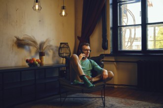 A man dressed in a green long-sleeve shirt and light pants relaxes in a chair, engaging in a phone