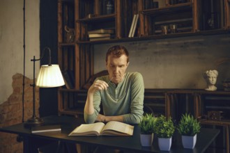 A man sits at a wooden desk in a warm, inviting study, deeply focused while reading a book. Shelves