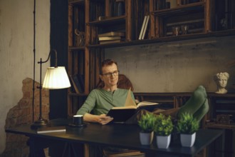 A middle-aged man relaxes with his feet up on a desk in a home office, reading a book. Natural
