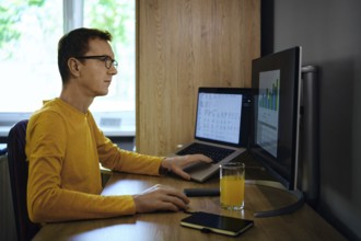A man wearing a yellow shirt sits focused at a desk, using a laptop and a monitor. He analyses data