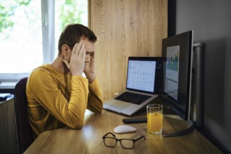 A middle-aged man sits at a wooden desk holding his head in frustration. Two computer screens