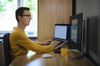 A middle-aged man focused on his work sits at a wooden desk with two computer screens. He enters