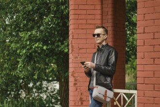 A man dressed in a leather jacket and sunglasses stands by red brick columns. He holds a smartphone