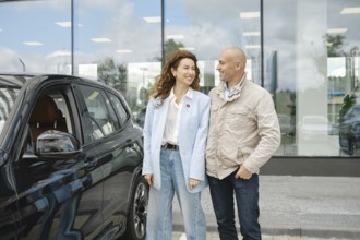 A couple stands beside a sleek black car at a bustling car dealership, engaged in a lively