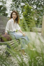 A woman with curly hair relaxes on a wooden bench in a serene park setting, wearing a white shirt