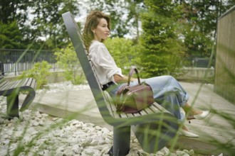 A woman sits comfortably on a bench in a park, surrounded by greenery and decorative stones. She