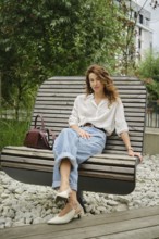 A woman sits comfortably on a unique wooden bench in a modern urban garden. She wears casual