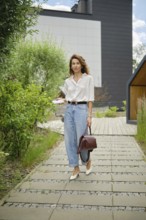 A woman walks gracefully along a garden path surrounded by lush greenery and modern architecture.