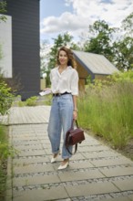 A fashionable woman strolls along a scenic pathway lined with lush plants. She wears a white shirt