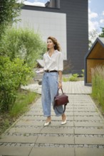 A woman wearing a white shirt and high-waisted jeans strolls through a landscaped path surrounded