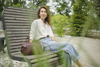 A woman with curly hair sits comfortably on a wooden bench in a lush park. Dressed in a light shirt