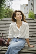A woman with curly hair sits relaxed on a wooden bench, wearing a white shirt and blue jeans. She