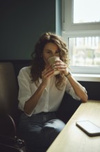 A woman with curly hair sits by a window in a cafe, holding a warm cup of coffee. Sunlight streams