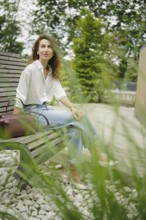A woman sits calmly on a wooden garden bench, dressed in a light shirt and jeans. Tall grass sways