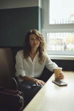 A woman with curly hair sits at a wooden table in a cafe, holding a paper cup. Sunlight streams