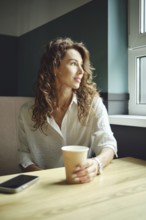 A woman with curly hair sits at a wooden table in a cozy cafe, holding a coffee cup. She gazes out
