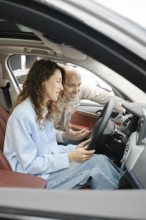 A woman sits in the driver's seat of a car, looking at the dashboard screen, while an older man