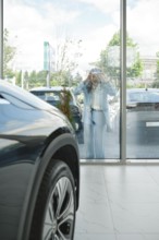 A woman stands outside looking in through large glass windows to a car dealership, admiring various