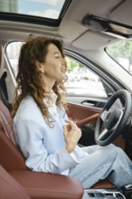 A woman with curly hair is seated in the driver's seat of a modern car, smiling and looking forward