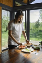 A woman arranges slices of salmon over tortilla on a wooden table in a bright kitchen. Sunlight
