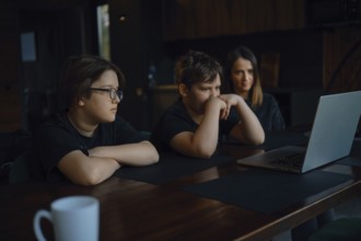 Two children and their mom watching movie on a laptop. They sit at a wooden table in a comfortable