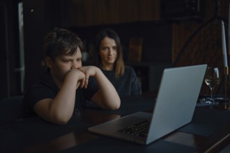 A boy sits focused at a table, watching a laptop screen closely while his mother looks on with a