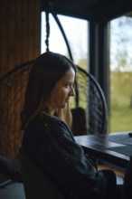 A woman is seated at a table in a warm, inviting indoor space with large windows. The setting is