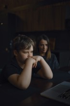 A young boy sits at a table, staring intently at a laptop screen with his hands on his chin. His