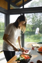 A woman is slicing beef meat in a sunlit kitchen with large windows. Fresh produce is spread on the