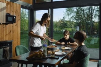 A mother serves a healthy meal to her two children at a stylish kitchen table surrounded by large