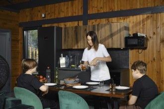 Two children sit at a dining table while their mother pours drink in a cozy cabin kitchen. A pan