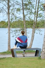A woman sits on a unique bench facing a serene lake, surrounded by trees. She enjoys the tranquil