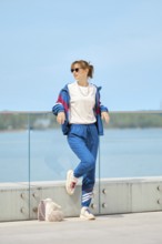 A woman relaxes by a glass railing at the waterfront on a bright sunny day. She wears a colorful