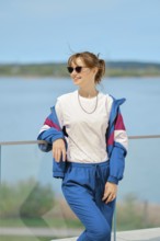 A woman stands confidently by the water, dressed in comfortable sportswear and sunglasses. She