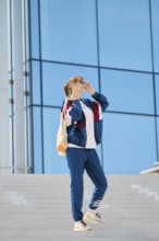 A young individual stands on a staircase, wearing sporty clothing and sunglasses. The sunny weather