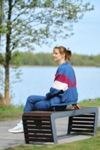 A young woman relaxes on a modern bench near a serene lake, dressed in colorful activewear. The