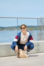 A young person squats on a modern deck by a scenic body of water, wearing sunglasses and a sporty