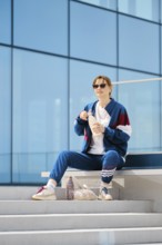 A young woman dresses in a stylish tracksuit sits on outdoor steps near a contemporary structure.