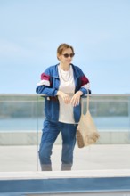 A woman stands by a glass railing at a waterfront location, dressed in a colorful jacket and