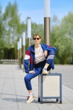 A woman dressed in a blue and maroon sporty outfit sits on a trashcan with one leg raised,