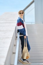 A young sporty woman with string bag stands on a staircase, dressed in stylish athletic track suit