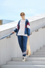 A young woman with sunglasses descends a staircase wearing a colorful sporty outfit. She carries a